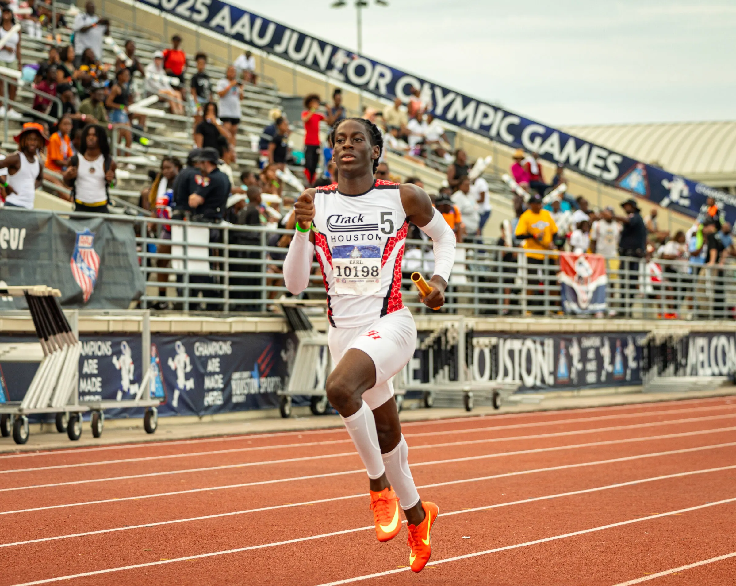A Track Houston athlete carries the baton down the home straight at the AAU Junior Olympic Games, grandstand roaring behind him.
